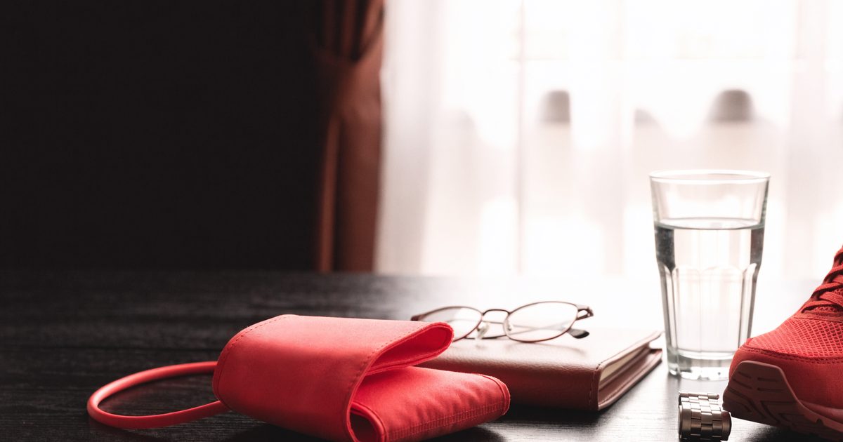 A red blood pressure cuff, reading glasses on a leather notebook, a glass of water, a wristwatch and a red running shoe on a dark wood table in soft window light.