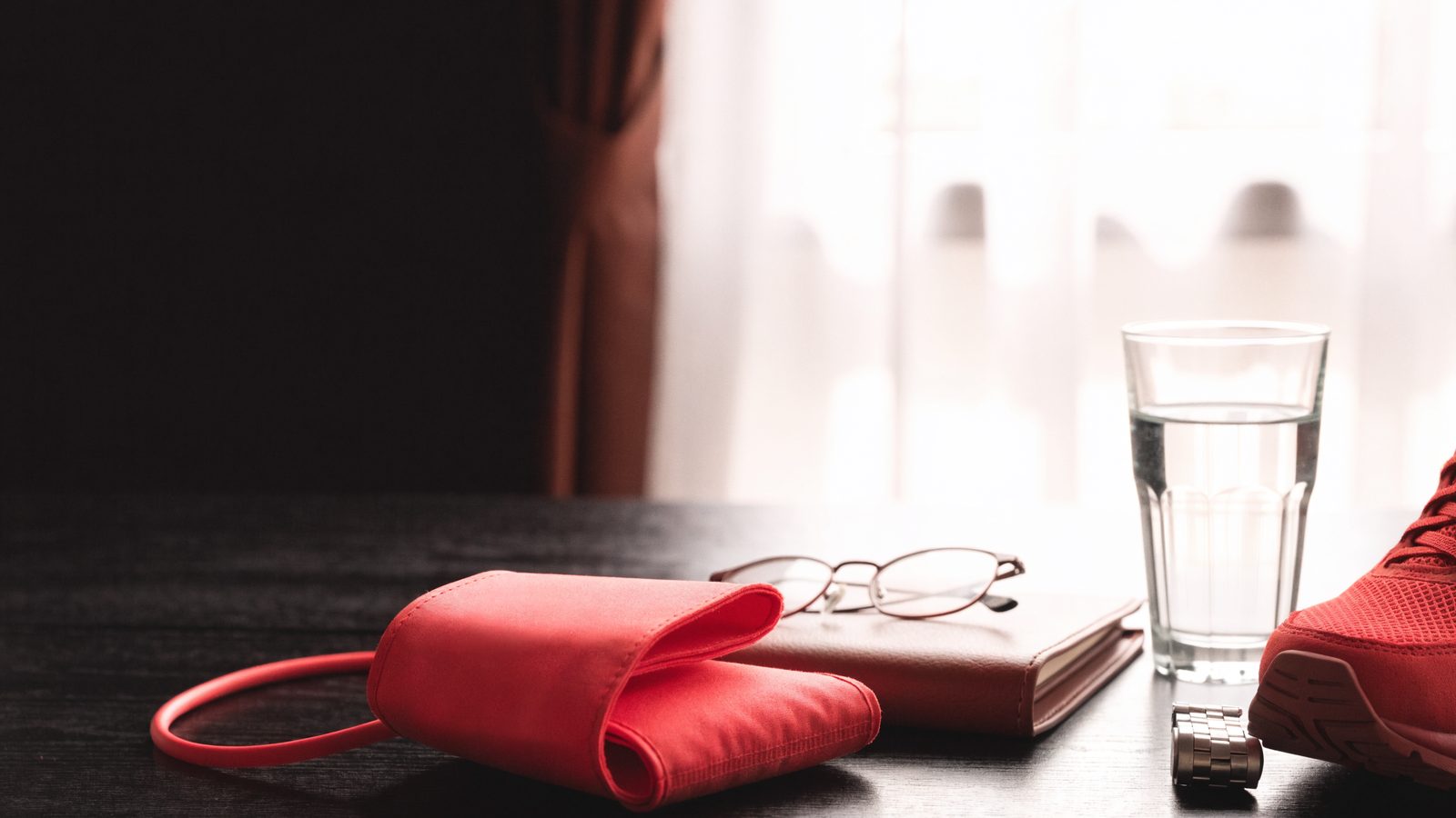 A red blood pressure cuff, reading glasses on a leather notebook, a glass of water, a wristwatch and a red running shoe on a dark wood table in soft window light.